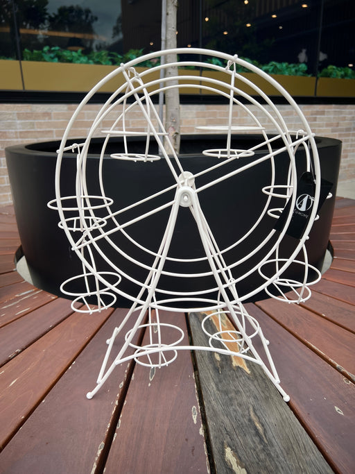 A white, ferris wheel-shaped dessert stand holding cupcakes, placed on a wooden surface with a green outdoor background.