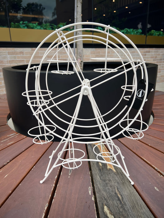 A white, ferris wheel-shaped dessert stand holding cupcakes, placed on a wooden surface with a green outdoor background.