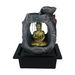 A Zen-inspired water feature with a bronze-colored meditating Buddha figure inside a rock-like structure, with water flowing over the figure in a black pot.